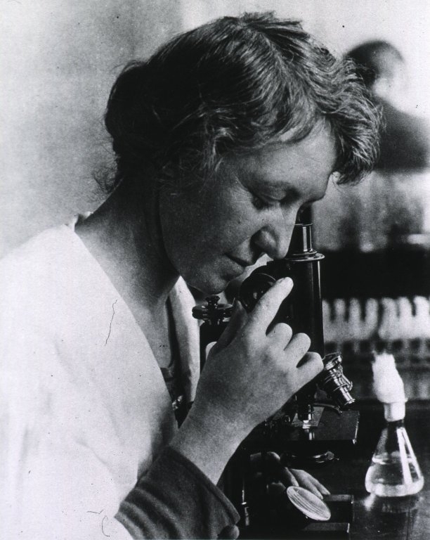 A black and white image of a woman looks into a microscope. The photo looks old fashioned.